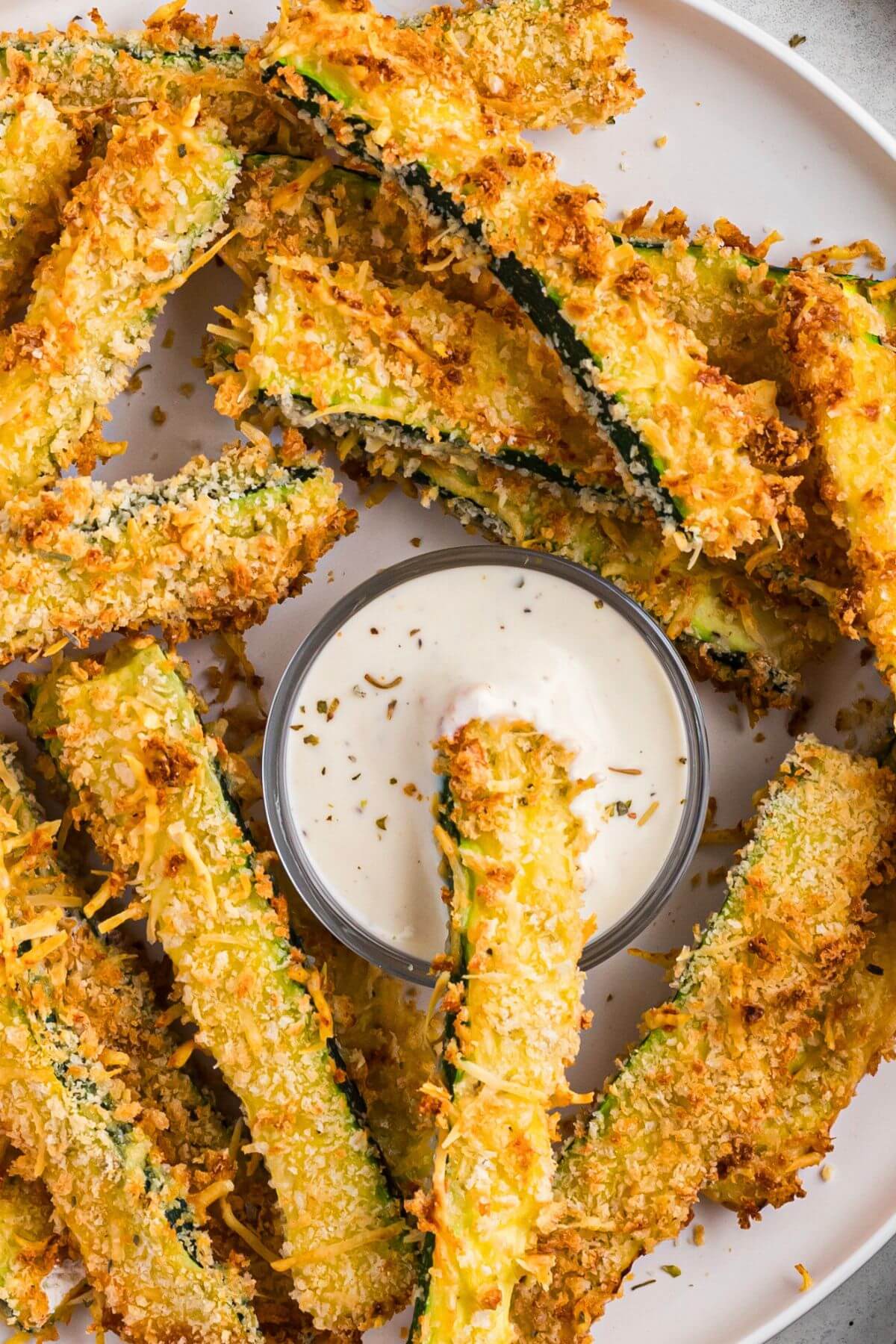 Golden crispy zucchini fries on a white plate with one fry in ranch dressing. 