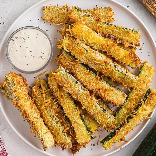 Golden crispy zucchini fries stacked on a white plate.