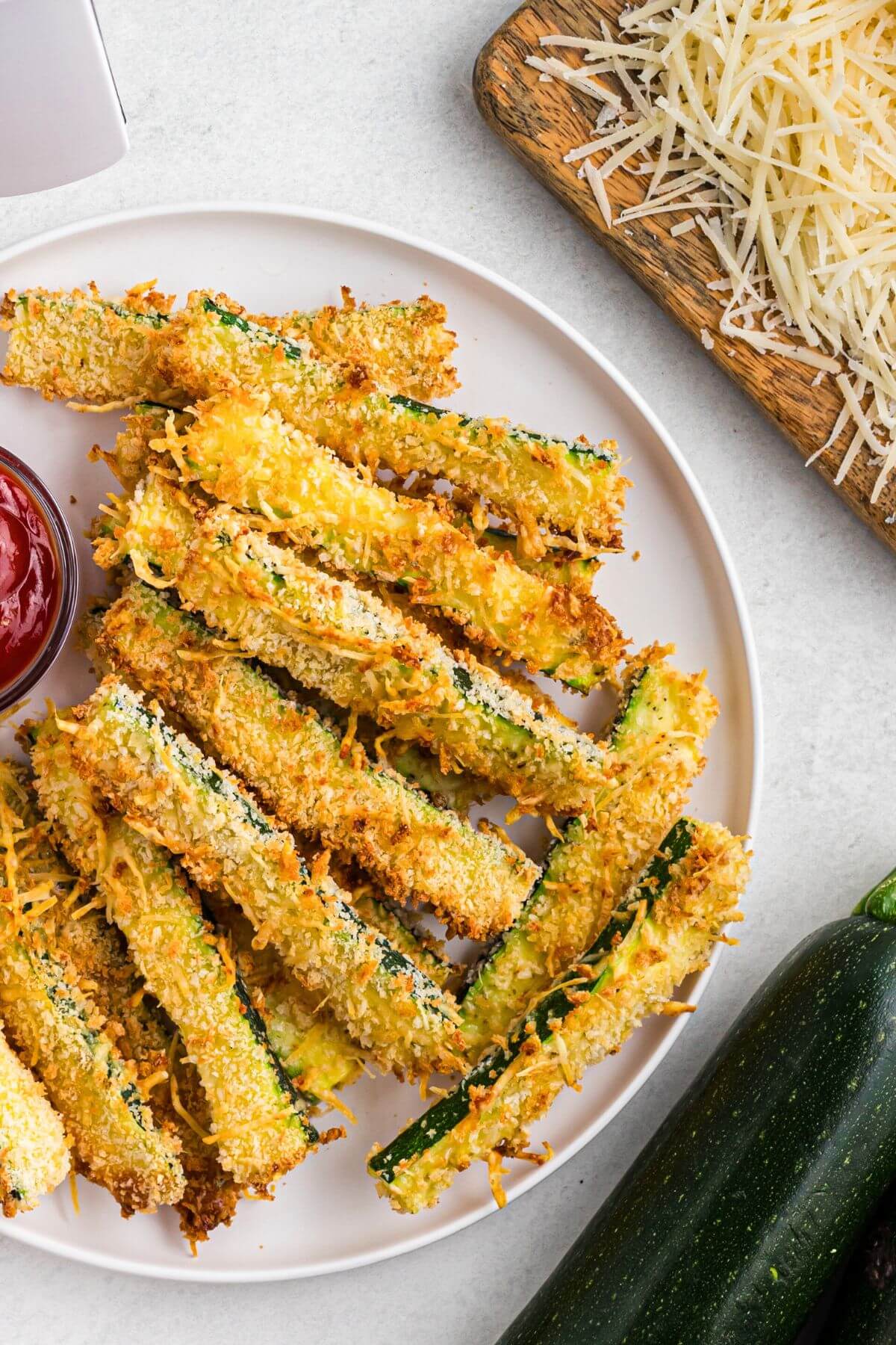 Golden crusted zucchini fries stacked on a white plate in front of the air fryer. 