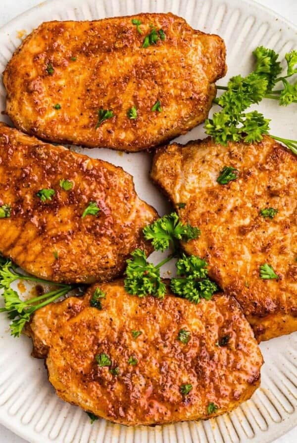 Golden juicy and seasoned pork chops on a white plate with parsley garnish.