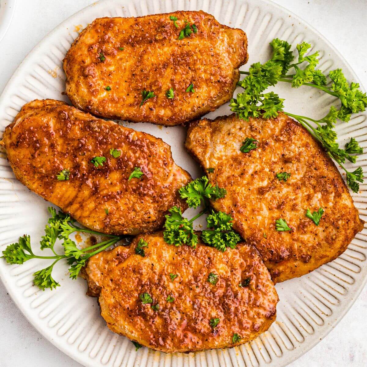 Golden juicy and seasoned pork chops on a white plate with parsley garnish.