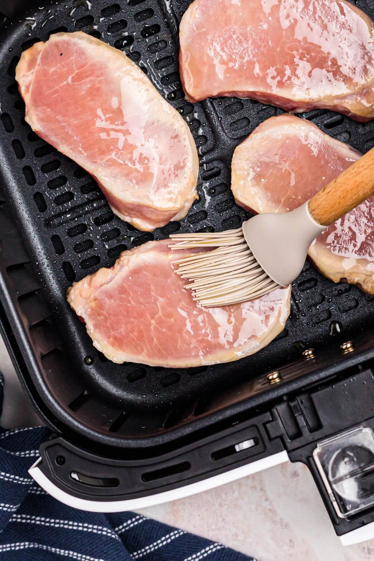 Uncooked pork chops in the air fryer basket being brushed with olive oil before being seasoned. 