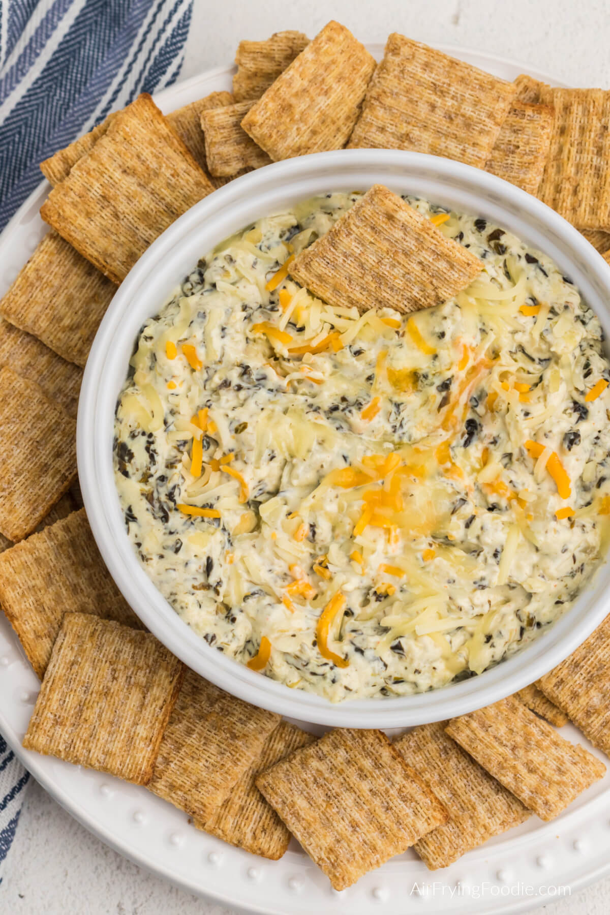 Air fryer artichoke dip topped with cheese and in an air fryer safe dish, surrounded by Triscuit crackers for dipping. 