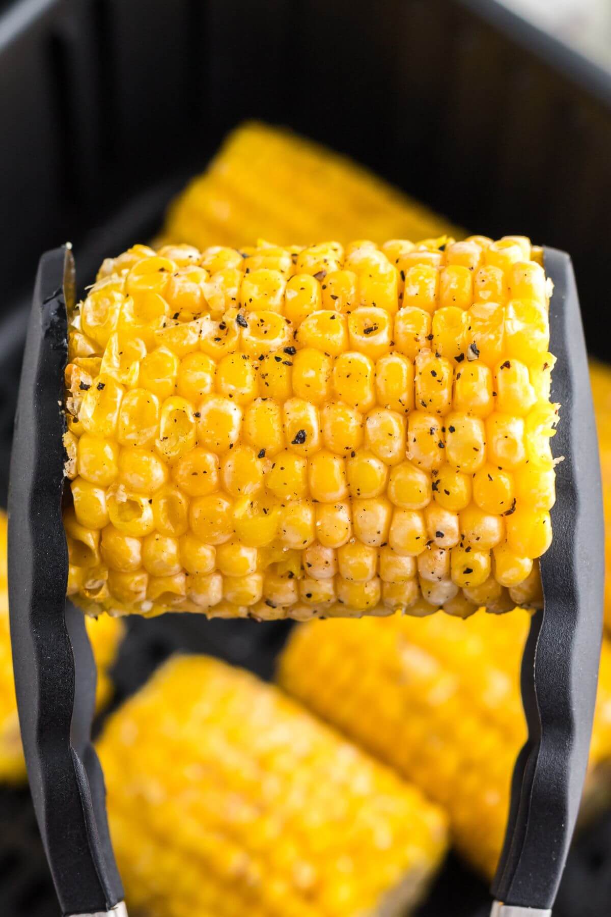 Cooked yellow corn on the cob being held with tongs and seasoned. 