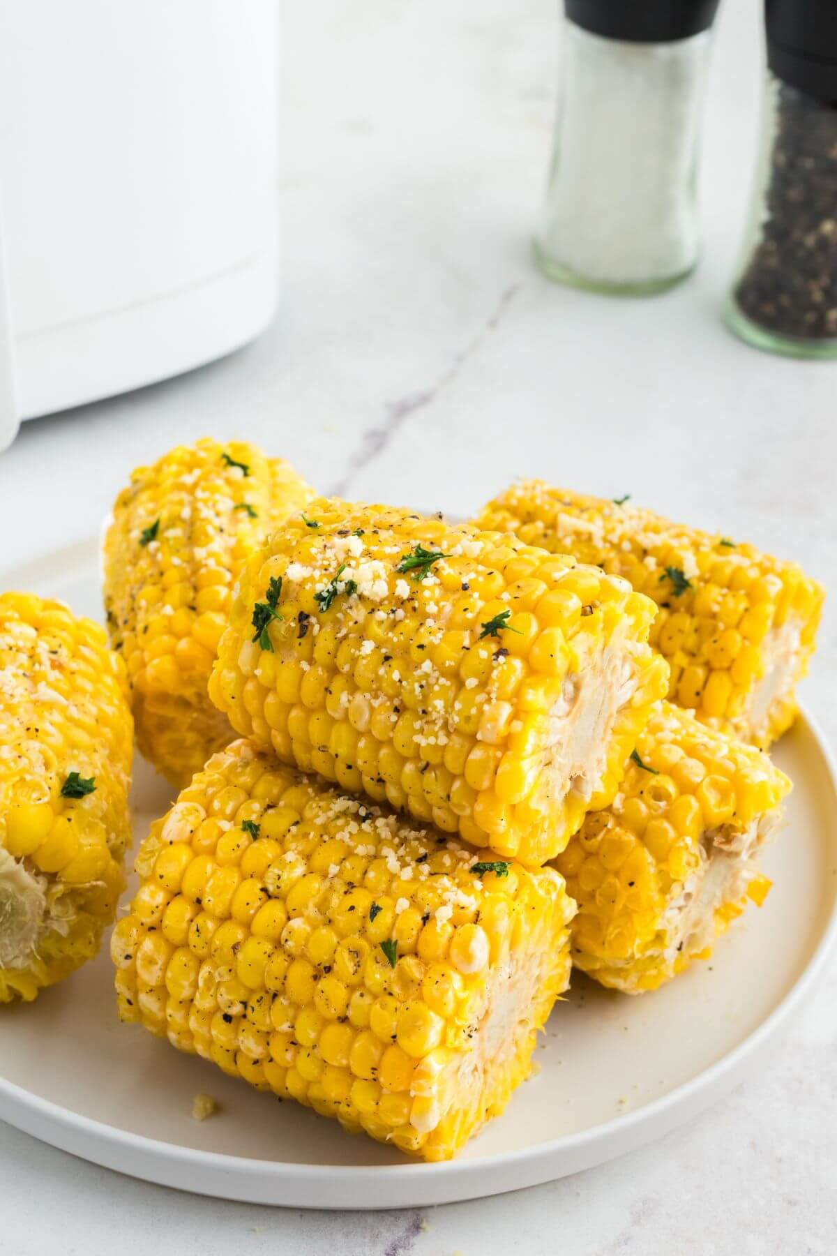 Golden yellow ears of corn stacked on a white plate garnished with parsley flakes. 