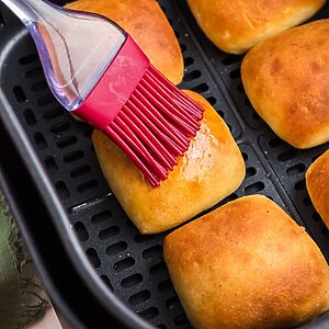 Golden brown dinner rolls being brushed with melted butter in the air fryer basket.