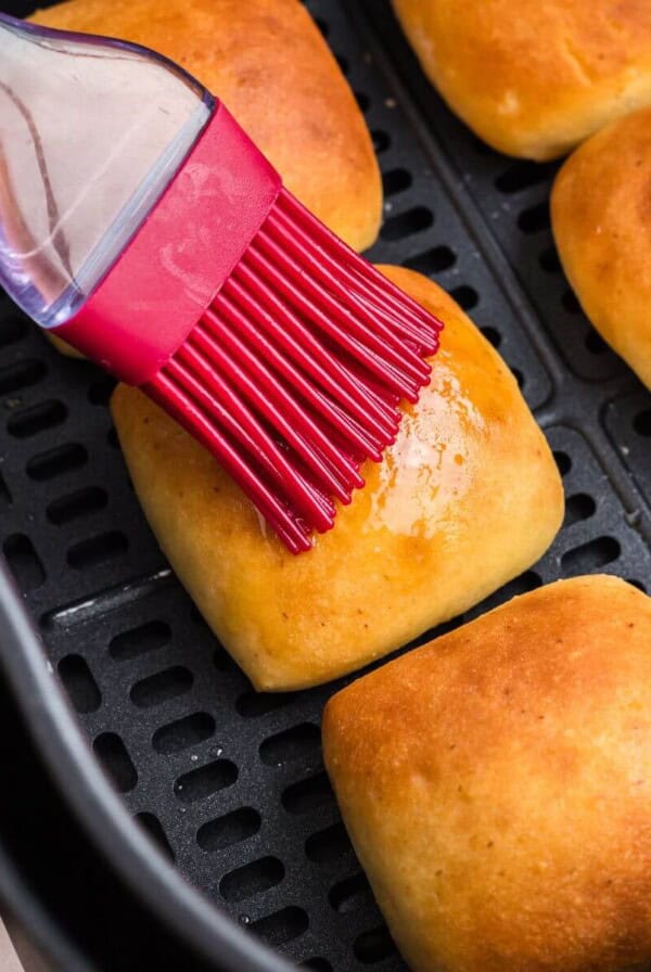 Golden brown dinner rolls being brushed with melted butter in the air fryer basket.