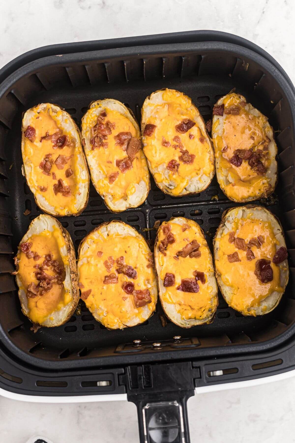 Baked potatoes stuffed with shredded melted cheese and chopped bacon, placed in the air fryer basket after being cooked. 