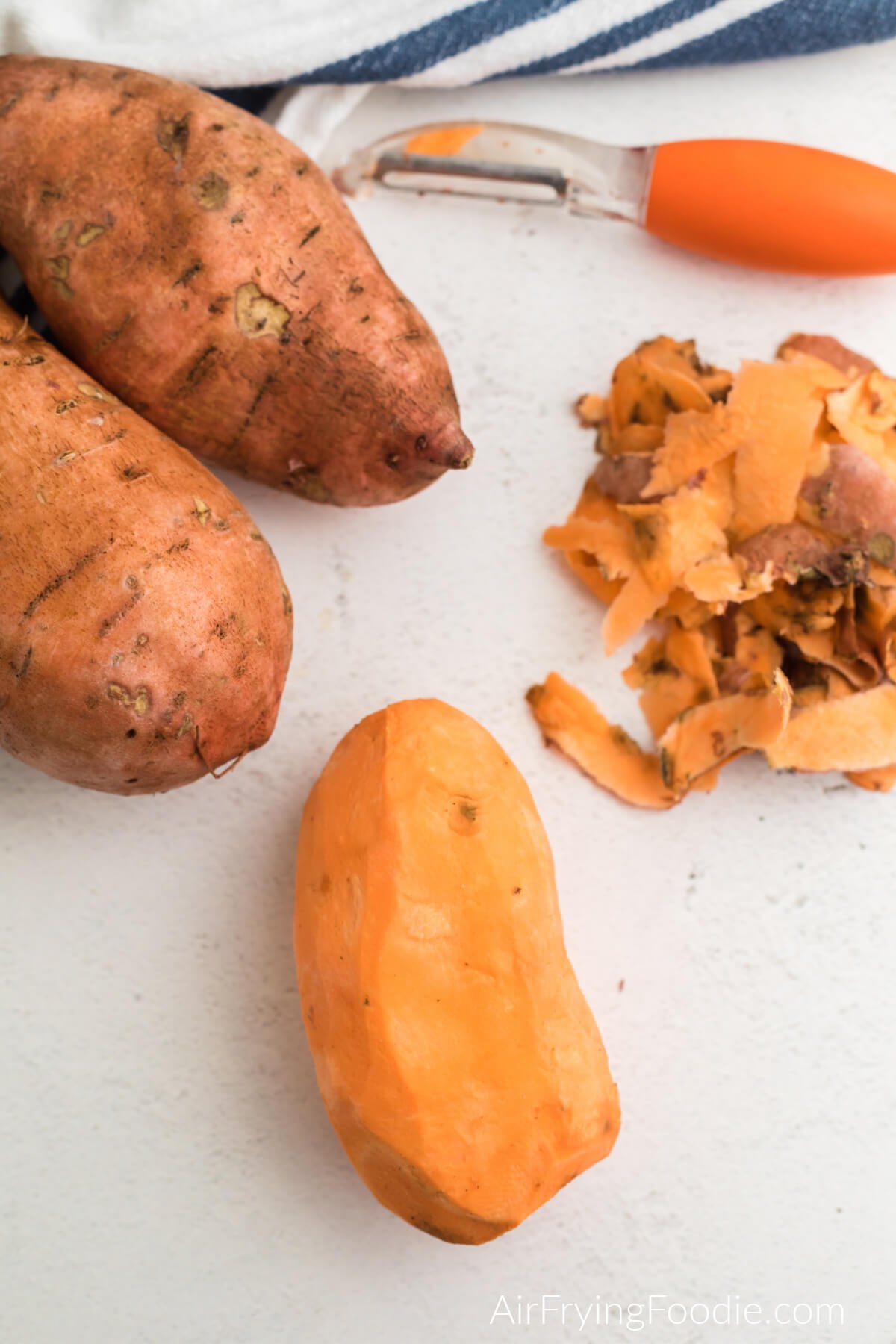 Peeled sweet potato on a table top surrounded by sweet potatoes and peelings with a vegetable peeler off to the side.