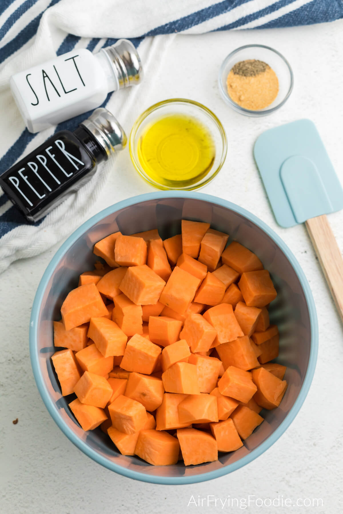 Sweet potato cubes in a mixing bowl with olive oil and seasoning and a spatular off to the side. 