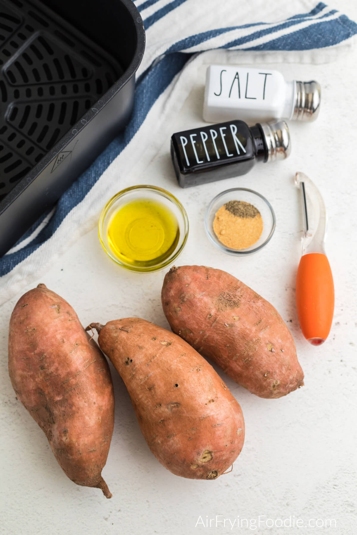 sweet potatoes, olive oil, garlic powder, pepper, an air fryer basket, and vegetable peeler on a white table.