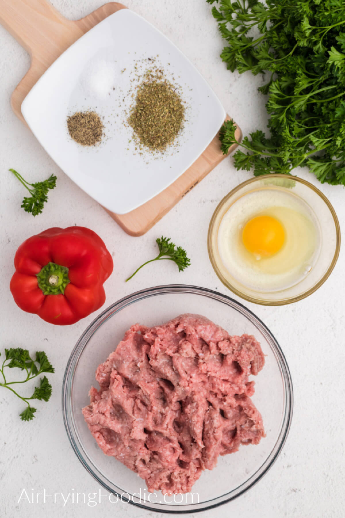Red bell pepper, ground turkey in a bowl, egg in a small bowl, and seasonings on a white plate, surrounded by fresh parsley. 