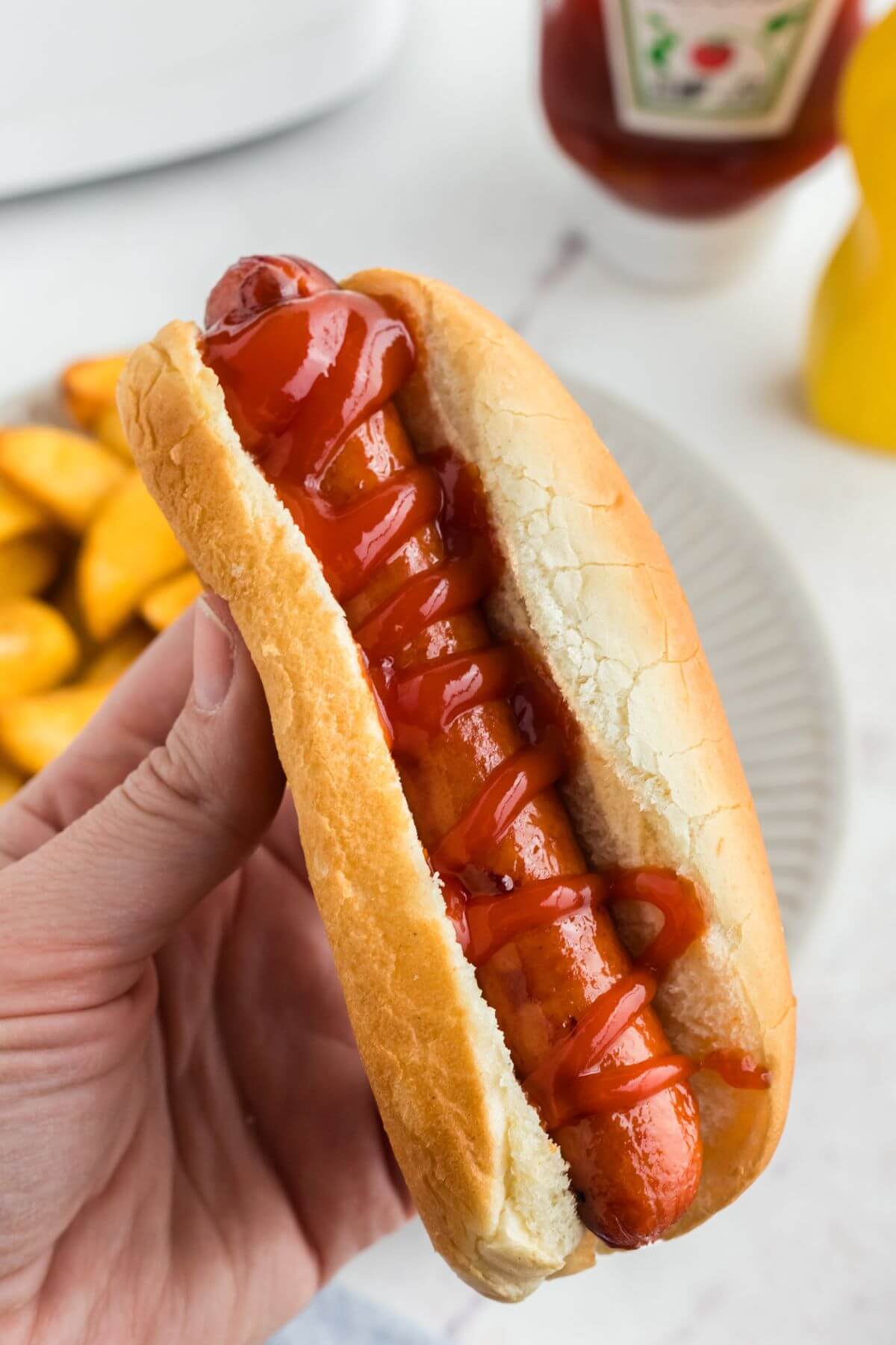Juicy golden brown hot dog in a bun with ketchup drizzled on top being held over a plate. 