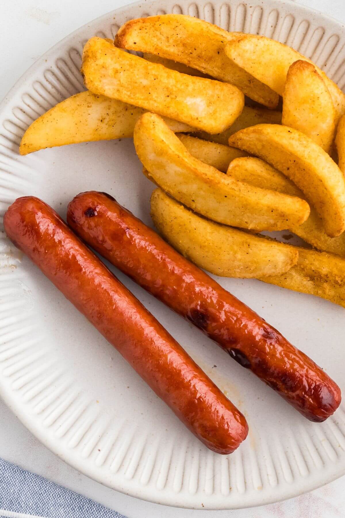 Juicy cooked hot dogs on a white plate with crispy french fries as a side dish. 