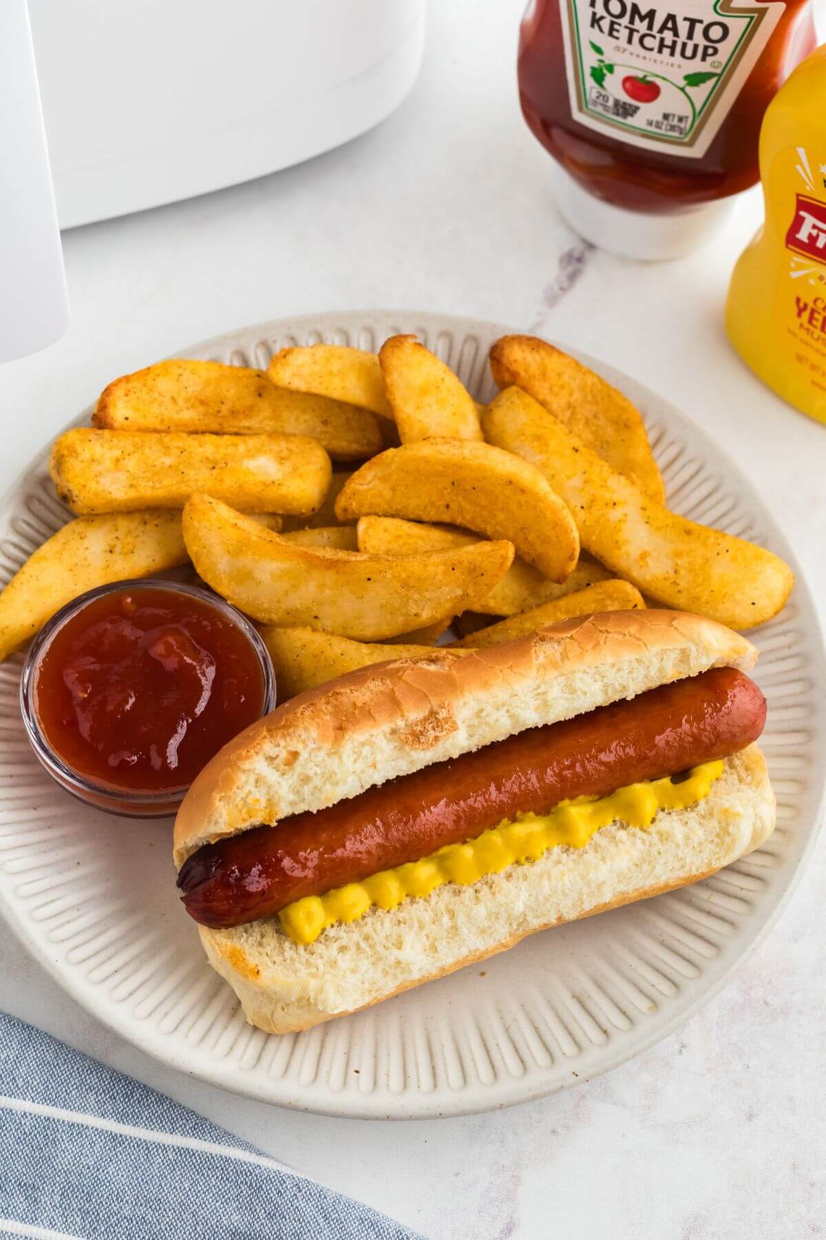 Juicy cooked hot dog in a bun with French fries on a white plate. 