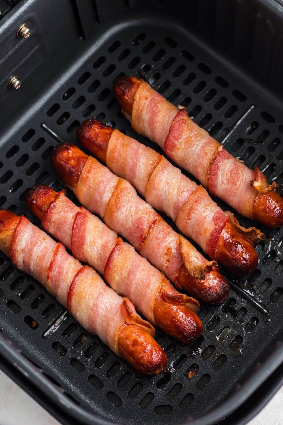 Juicy golden brown hot dogs wrapped in cooked bacon in the air fryer basket. 