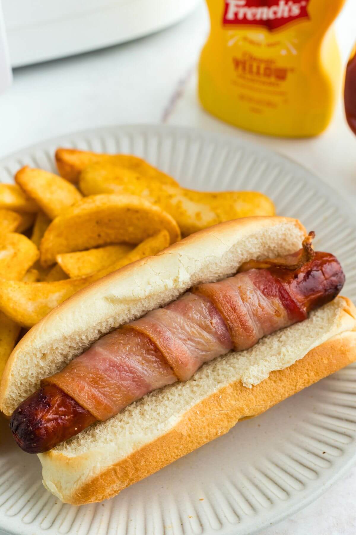 Juicy bacon wrapped hot dog in a bun with steak fries on a white plate. 
