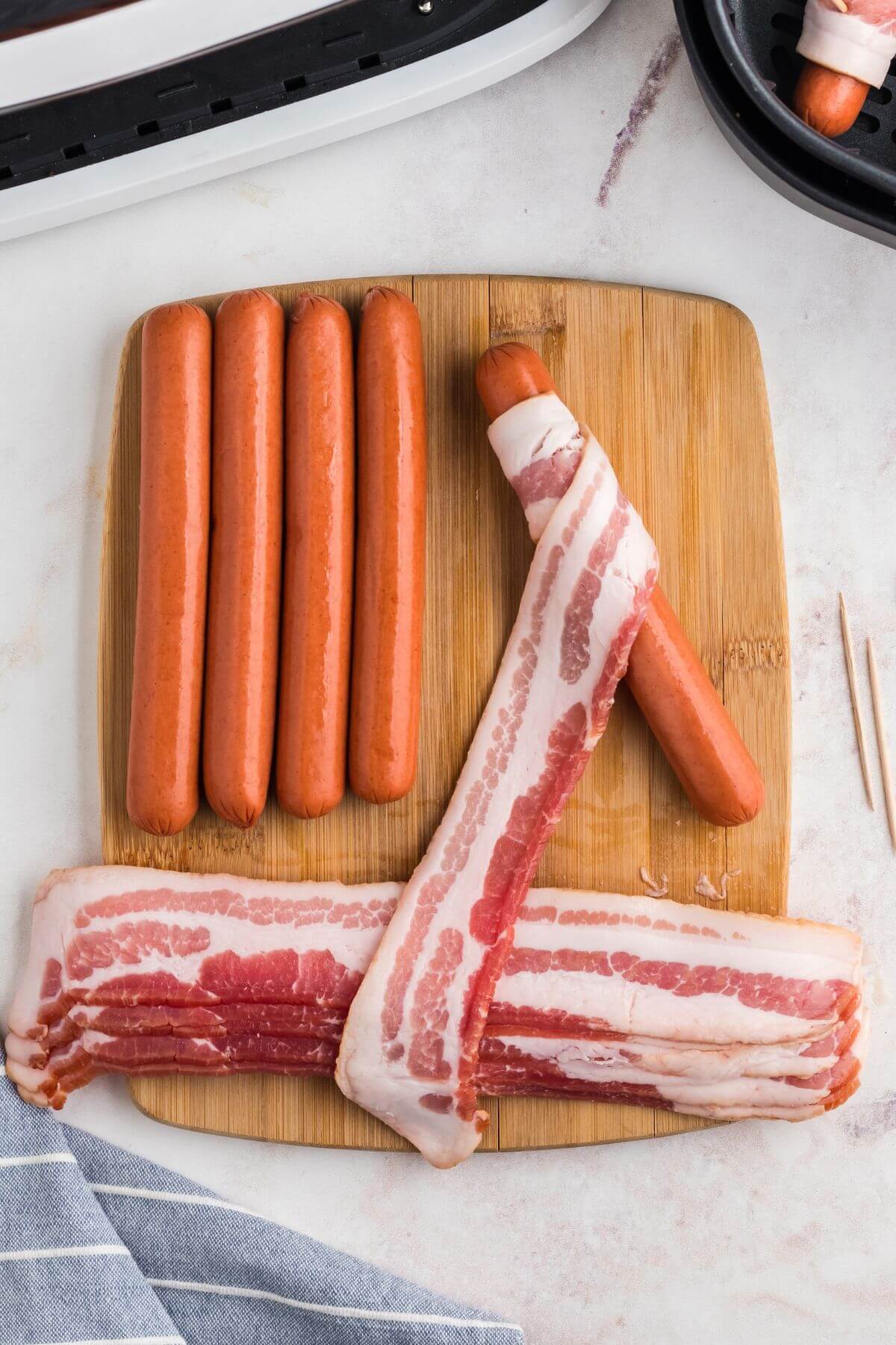 Strip of uncooked bacon wrapped around uncooked hot dog on a wooden cutting board. 