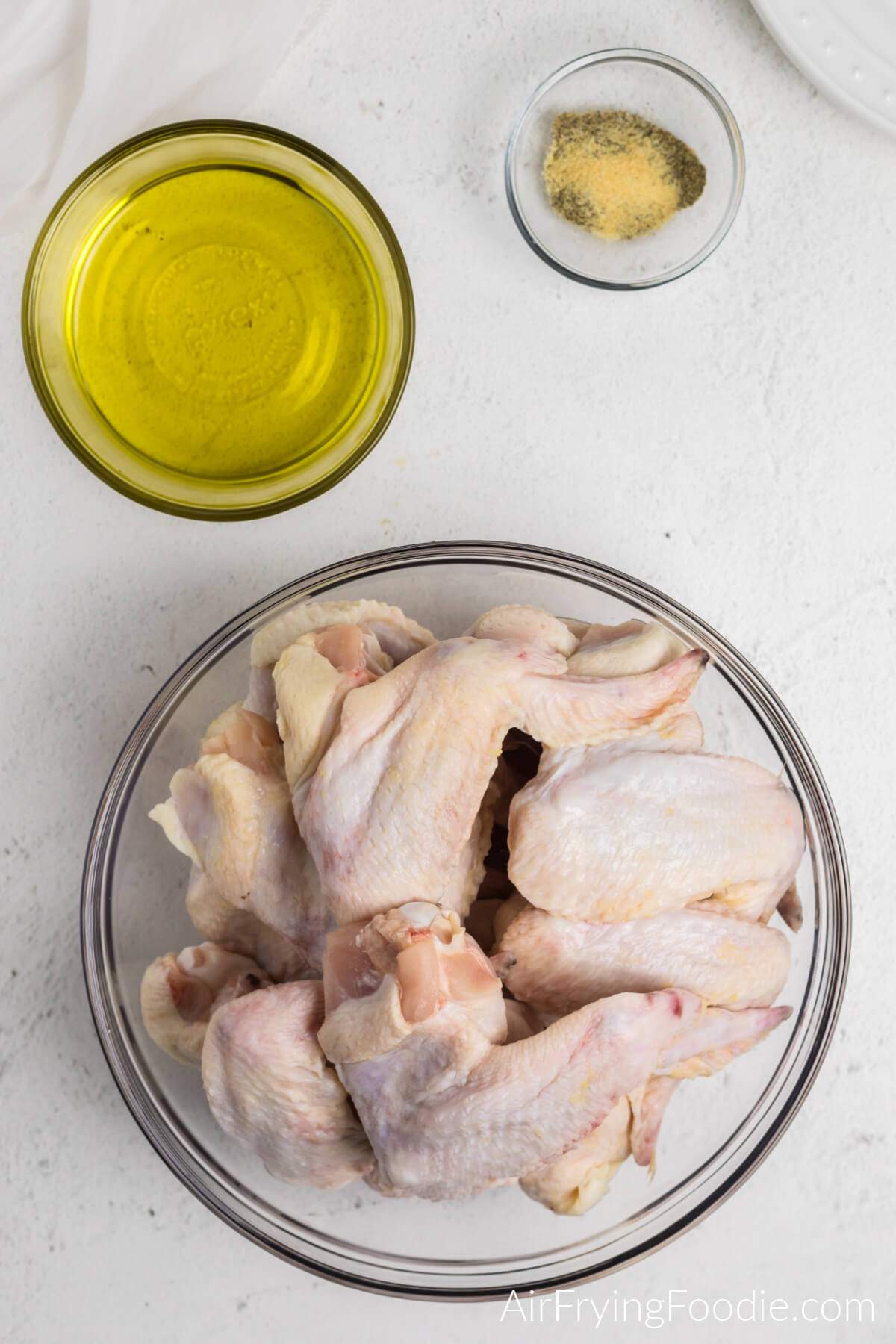 Ingredients needed to make homemade chicken wings in the air fryer: whole chicken wings in a bowl, a bowl of olive oil, and a bowl with black pepper, onion powder, and garlic powder. 