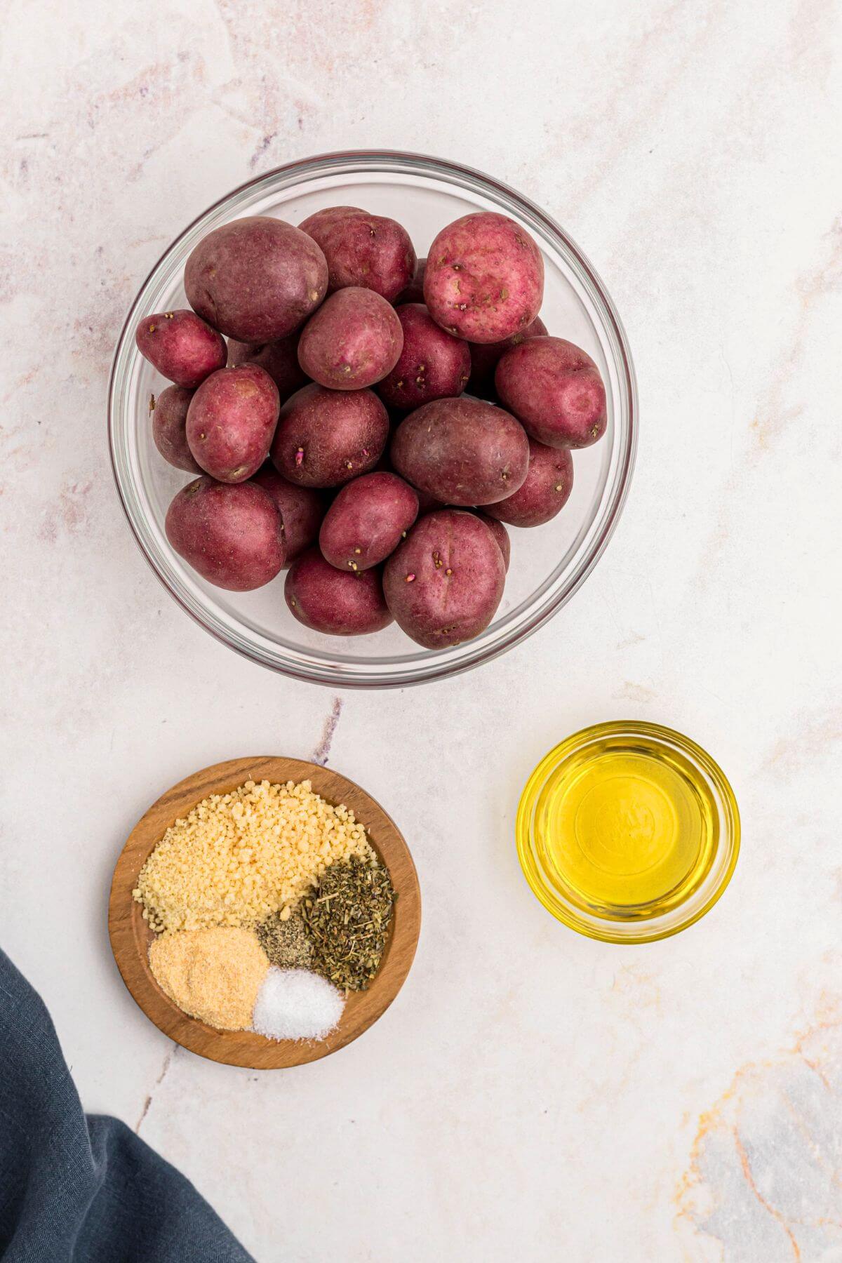 Uncooked red baby potatoes in a clear glass bowl with olive oil, seasonings and parmesan cheese on a marble table.