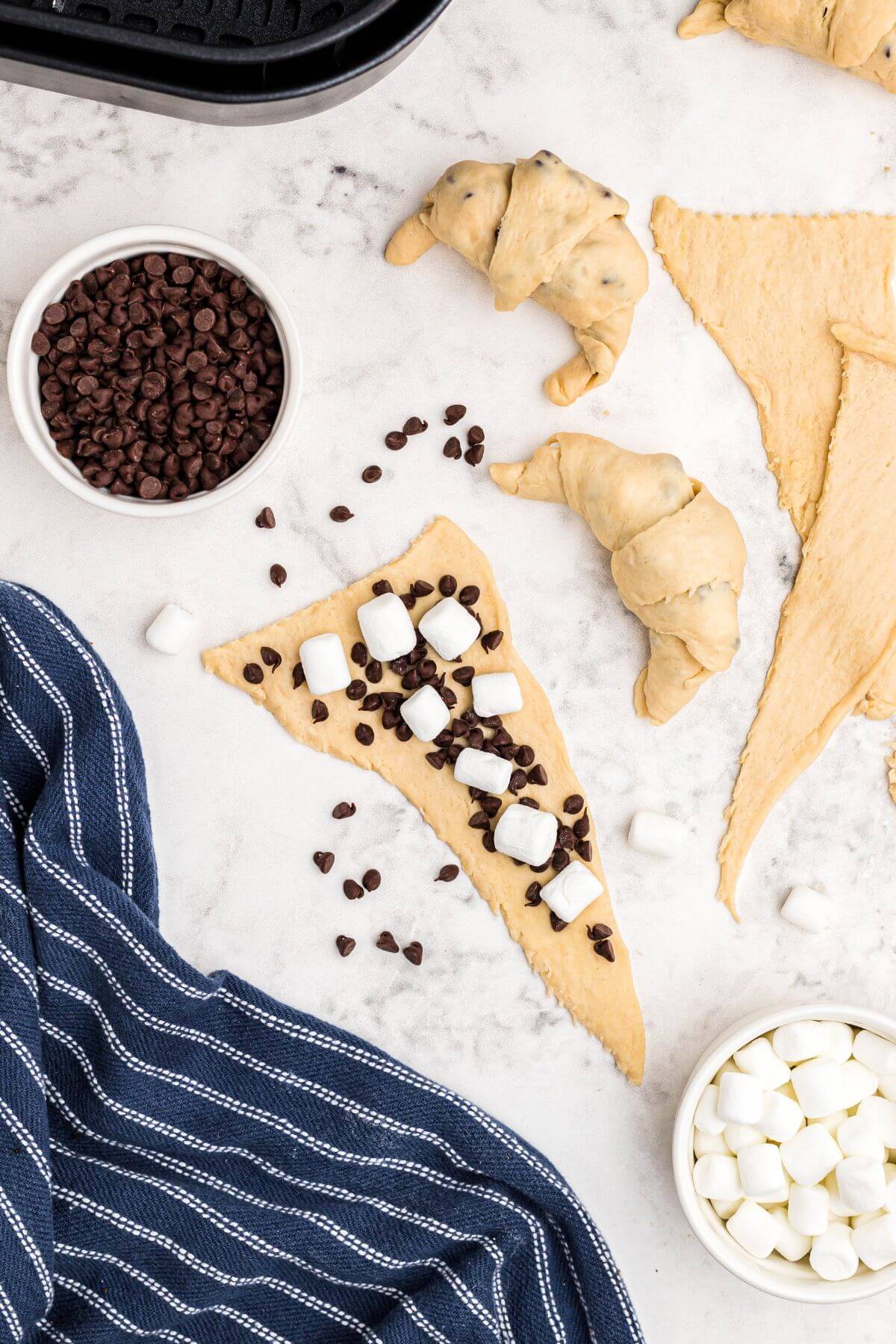 Unrolled crescent rolls topped with mini chocolate chips and mini marshmallows on a marble table being rolled into crescent shapes. 