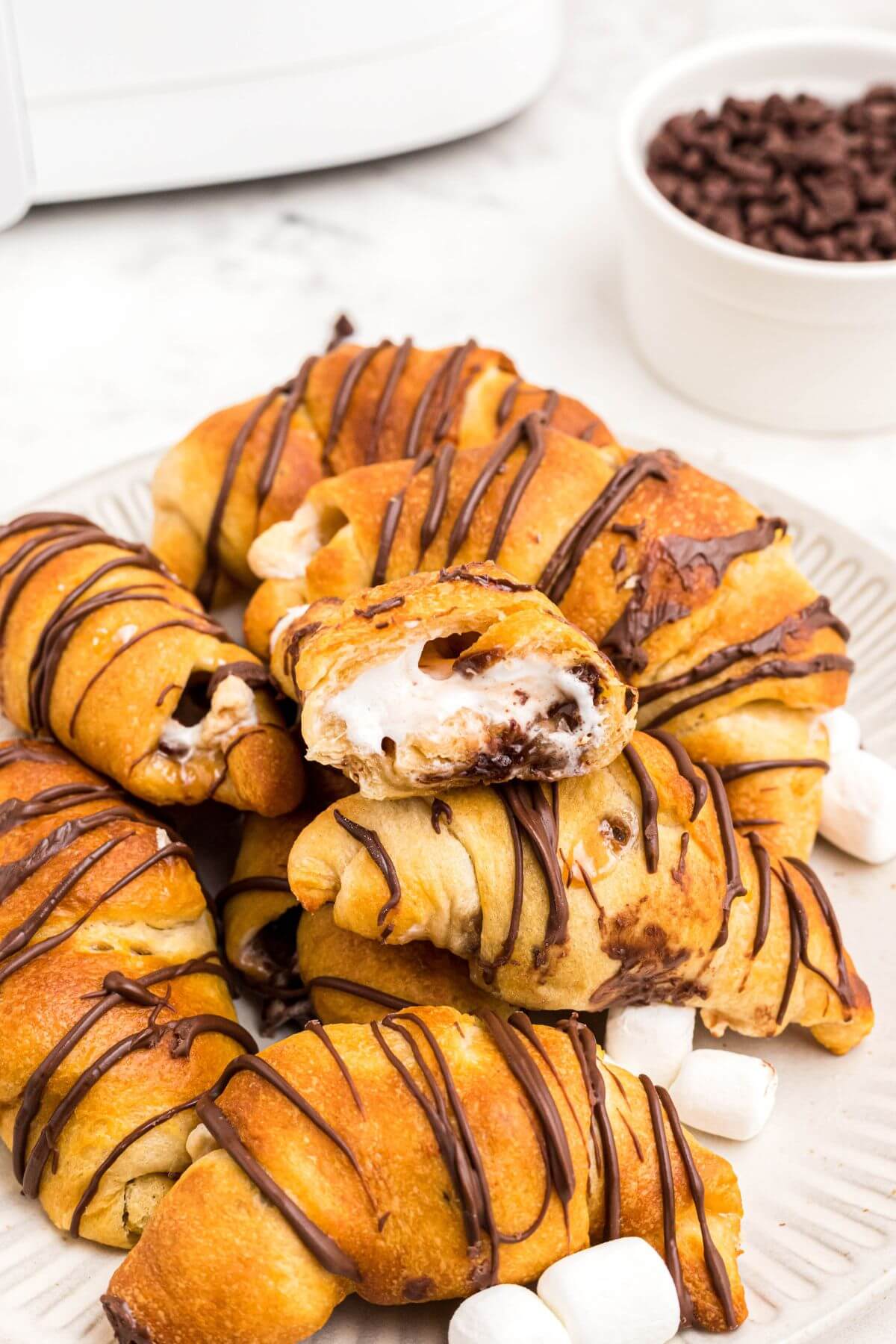 Golden flaky crescent rolls filled with gooey marshmallows and melty chocolate chips stacked on a plate with mini marshmallows and chips on the plate. 