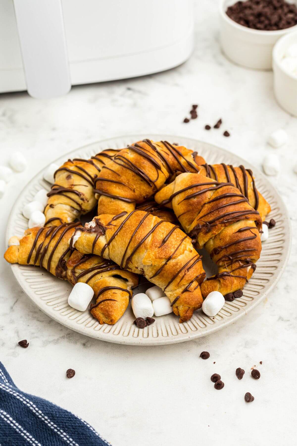 Golden flaky crescent rolls filled with marshmallows and chocolate chips, stacked on a cream plate with a blue linen. 