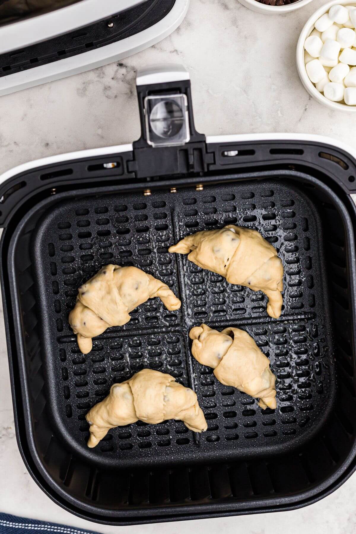 Uncooked rolled crescent rolls filled with mini chocolate chips and mini marshmallows, in an air fryer basket. 