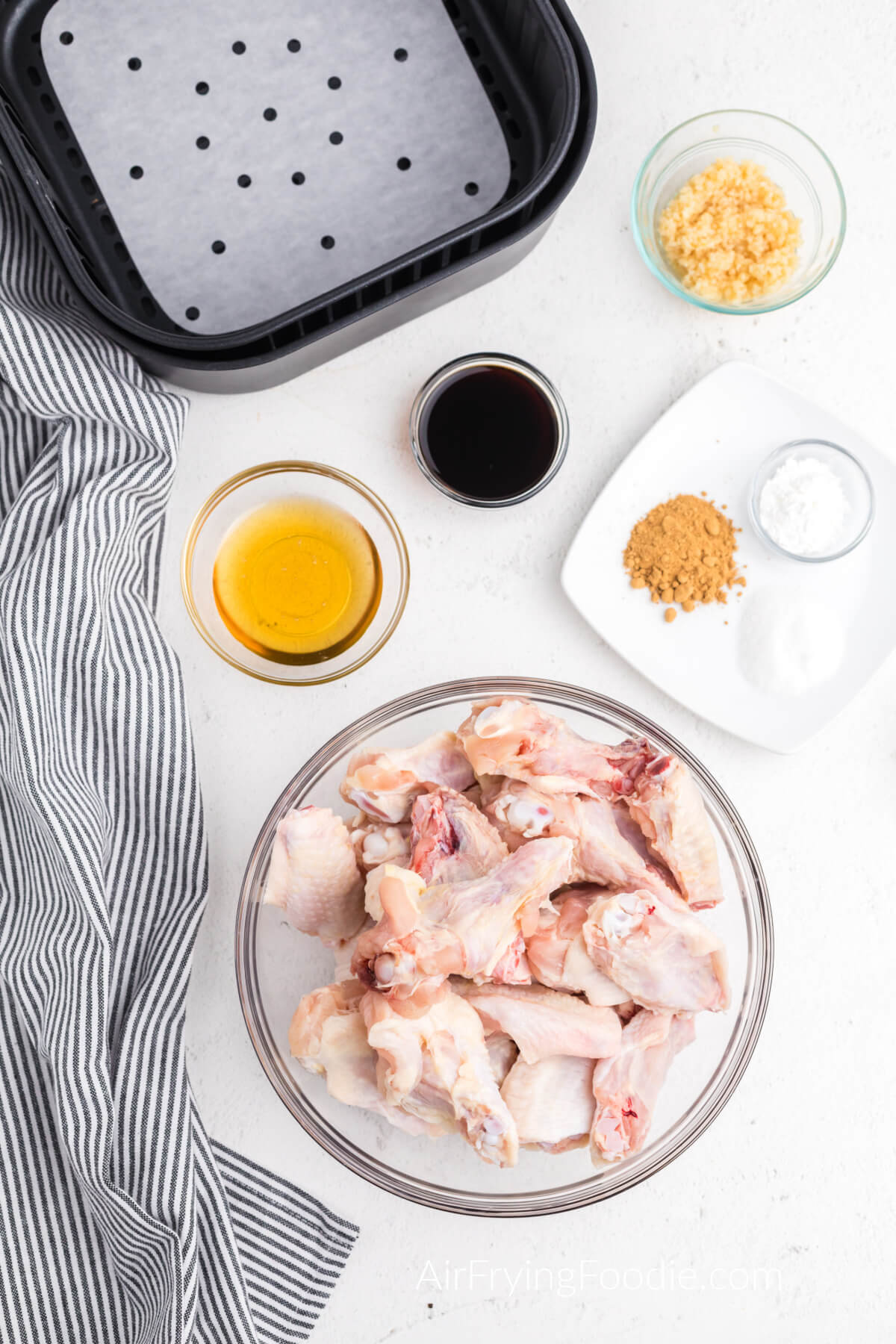 Chicken wings, honey, garlic, soy sauce, and other ingredients on a white plate next to a prepared air fryer basket. 