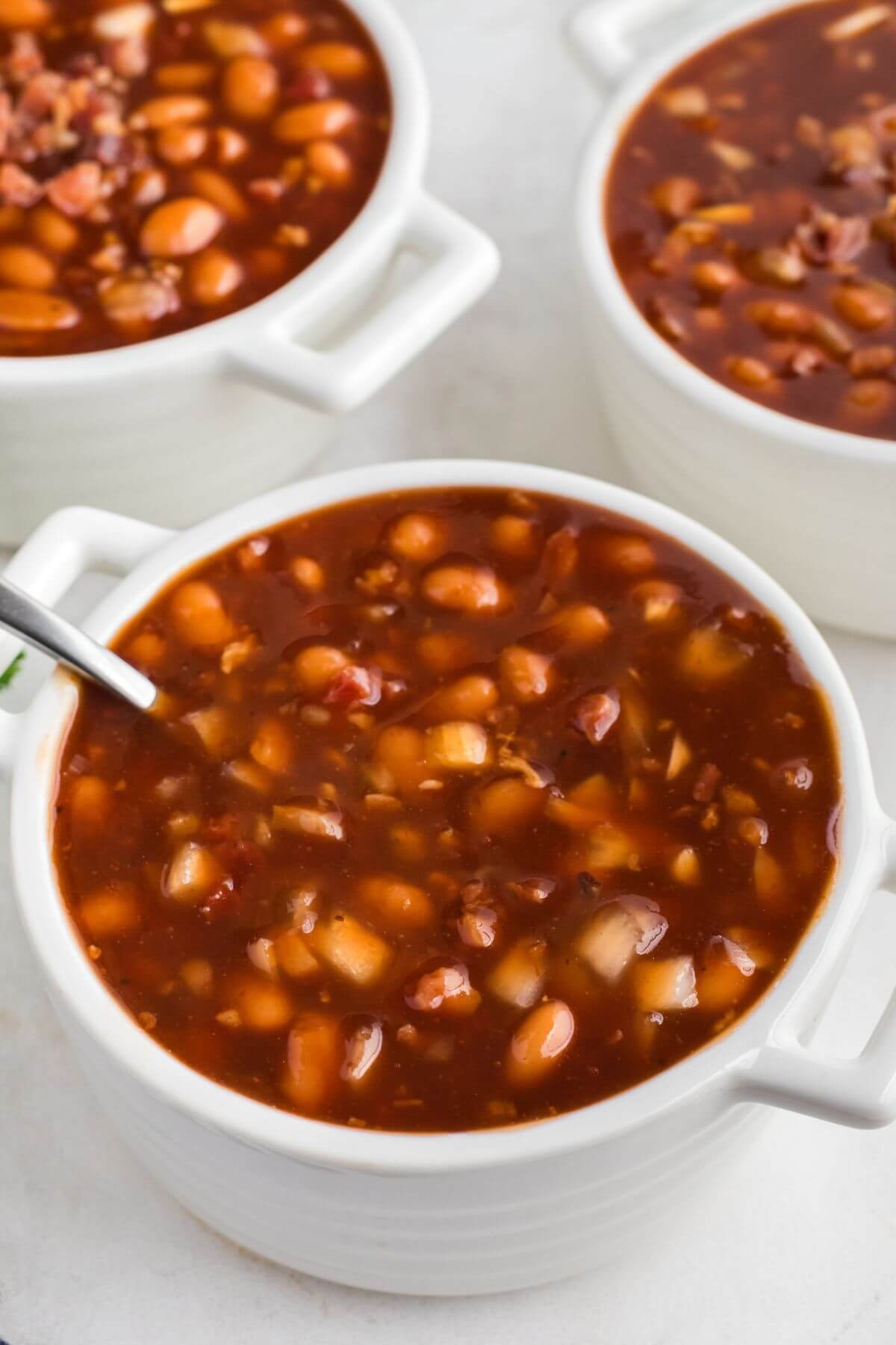 Juicy brown baked beans in white bowls with a spoon on the side. 