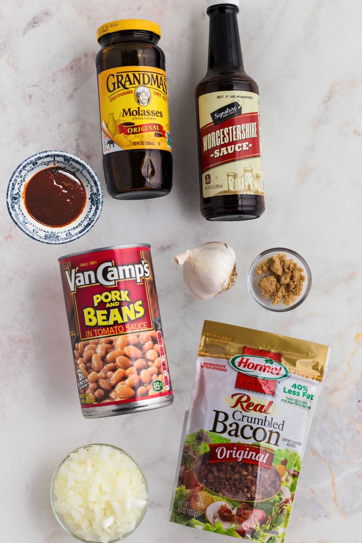 Ingredients of molasses, pork and beans, chopped onions, crumbled bacon, and more, needed to make baked beans from scratch on a marble table. 