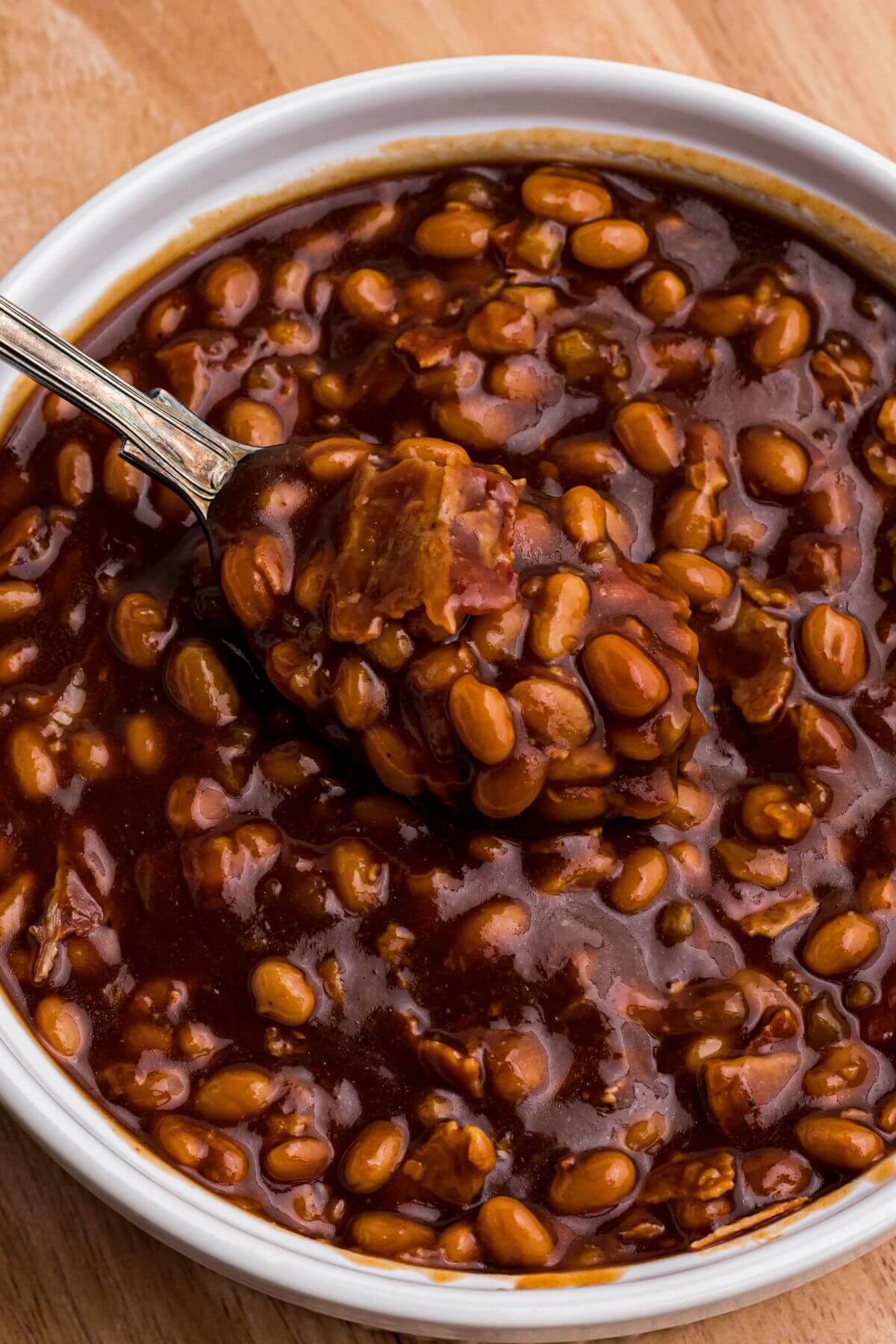 Juicy golden brown baked beans being scooped up with a spoon topped with bacon crumbles. 