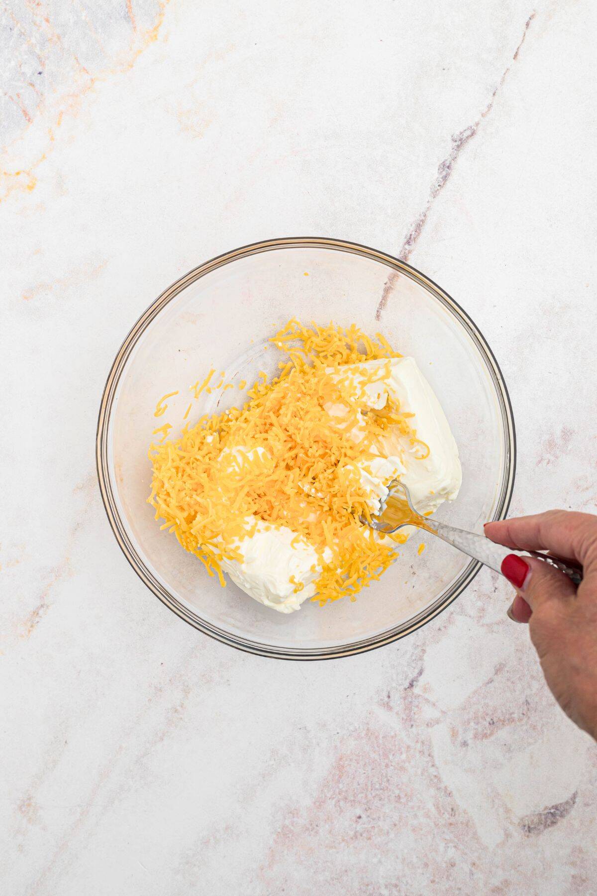 In a medium clear glass bowl, shredded cheese and cream cheese are being mixed together with a fork. 