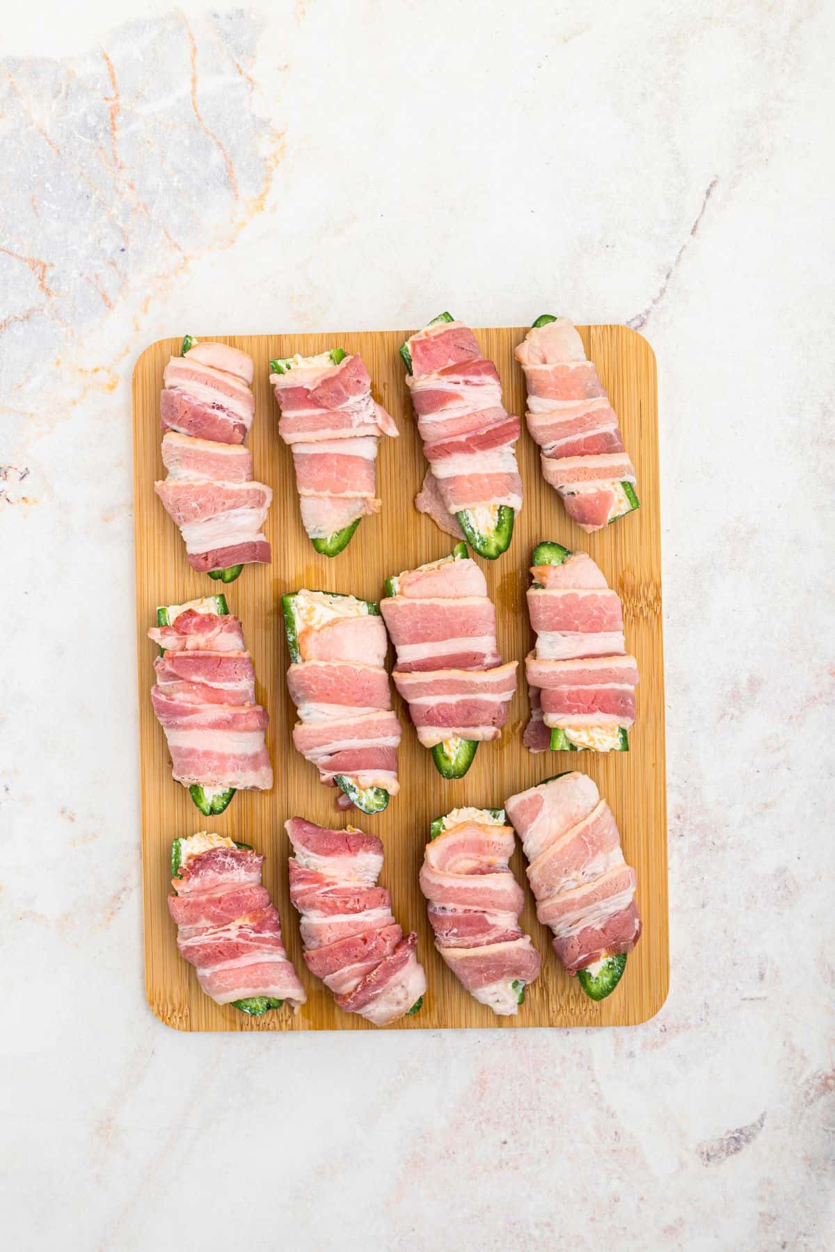 Cheese stuffed jalapenos, wrapped in slices of bacon, on a wooden cutting board. 