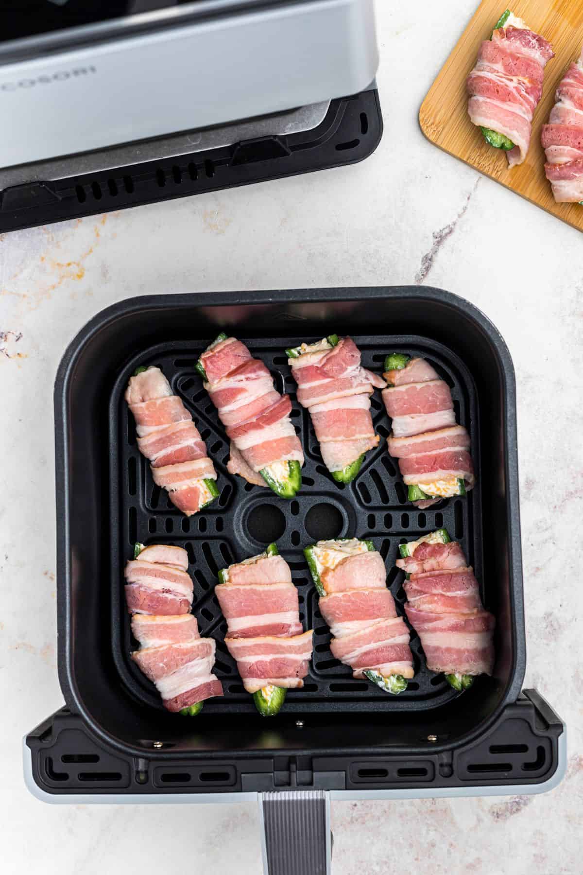 Uncooked bacon wrapped jalapenos in the air fryer basket, in front of the air fryer. 