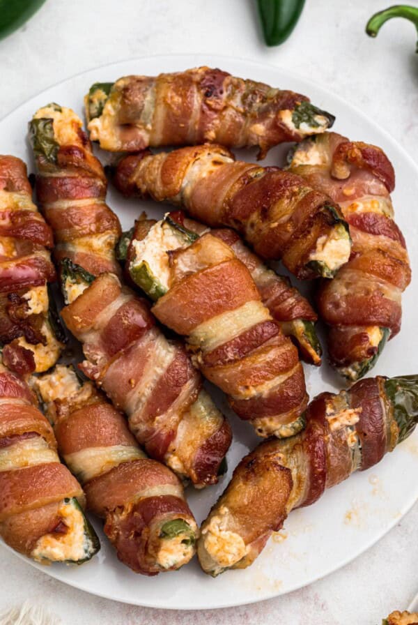 Crispy cooked bacon wrapped jalapenos, filled with cheese mixture, on a white plate.