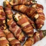 Crispy cooked bacon wrapped jalapenos, filled with cheese mixture, on a white plate.