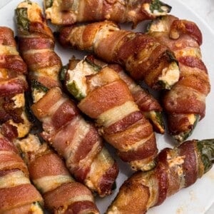 Crispy cooked bacon wrapped jalapenos, filled with cheese mixture, on a white plate.
