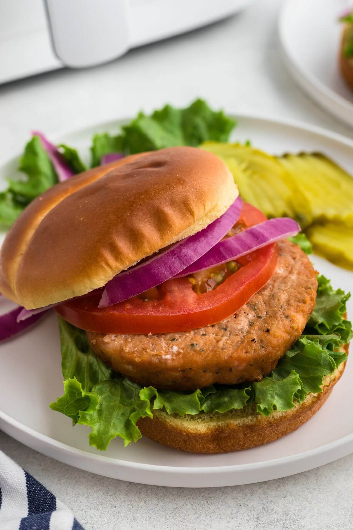 Salmon burger on a hamburger bun with lettuce, onion, tomato, and pickles on the side. 
