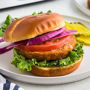 Salmon burger on a bun with lettuce, tomato, and sliced onion on a white plate.