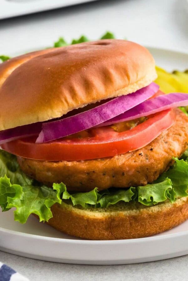 Salmon burger on a bun with lettuce, tomato, and sliced onion on a white plate.