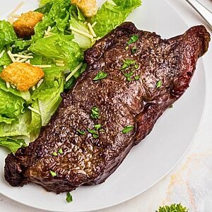 Juicy New York strip steak on a white plate with a salad.