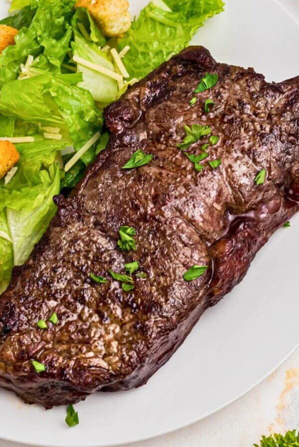 Juicy New York strip steak on a white plate with a salad.