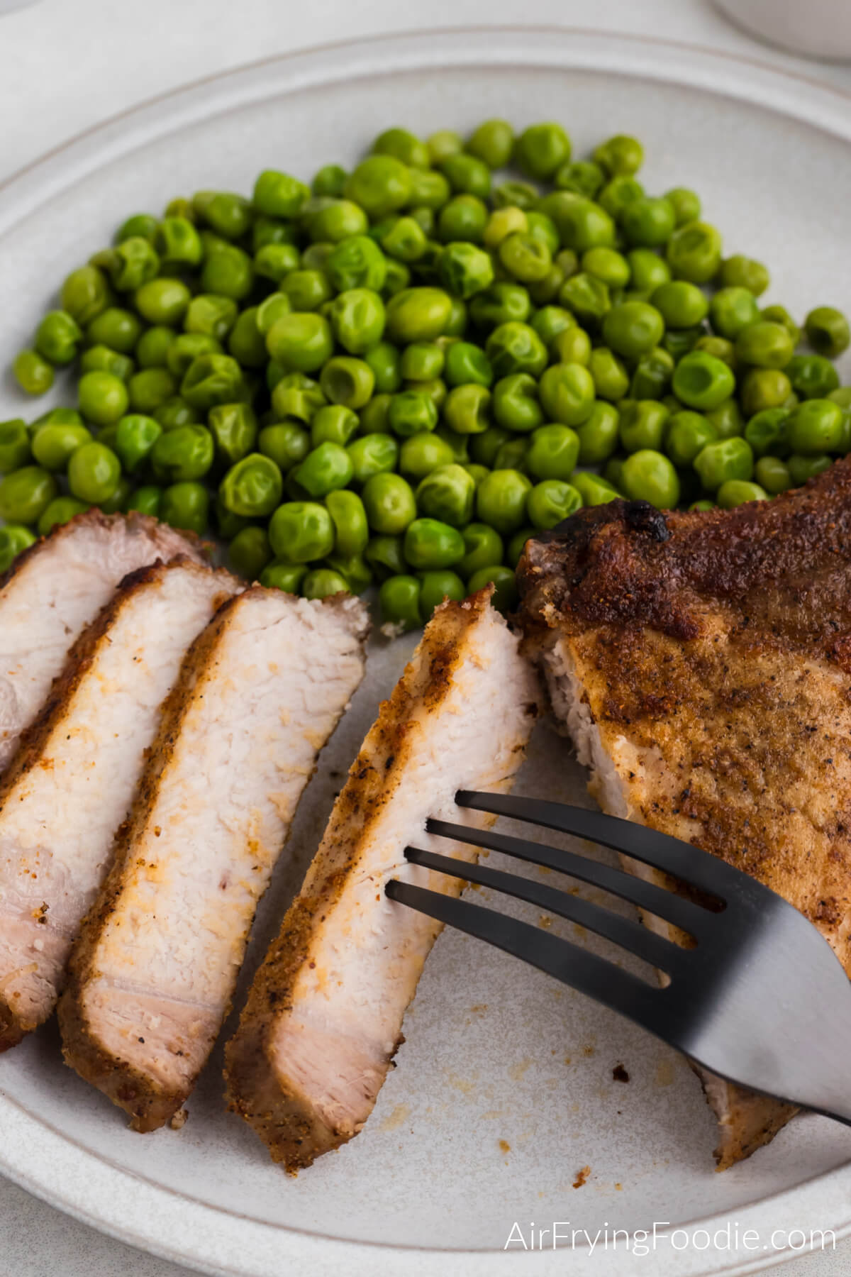Sliced pork steak on a white plate with a side of cooked green peas.