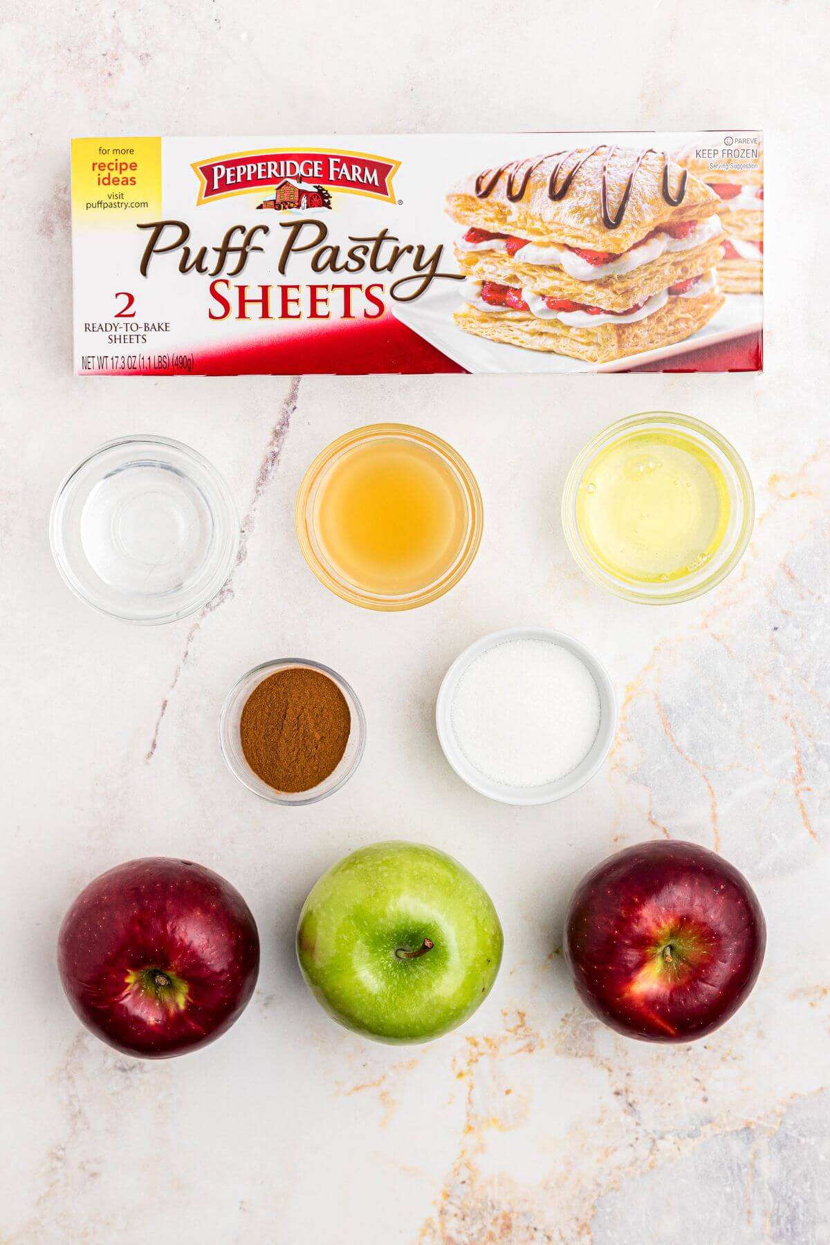 Apples, puff pastry, and other ingredients needed to make apple rings on a marble table. 
