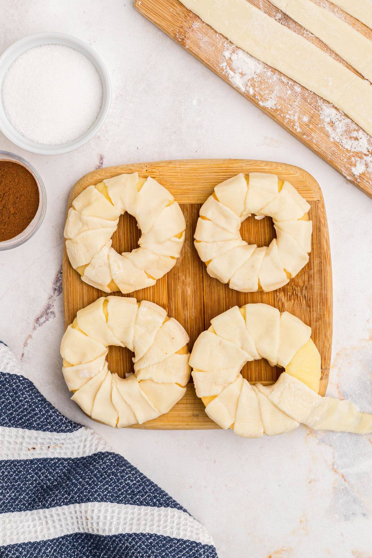 Apple rings being wrapped with strips of puff pastry on a wooden cutting board. 