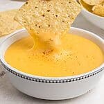 Tortilla chip being dipped into melted cheese in small white bowl.