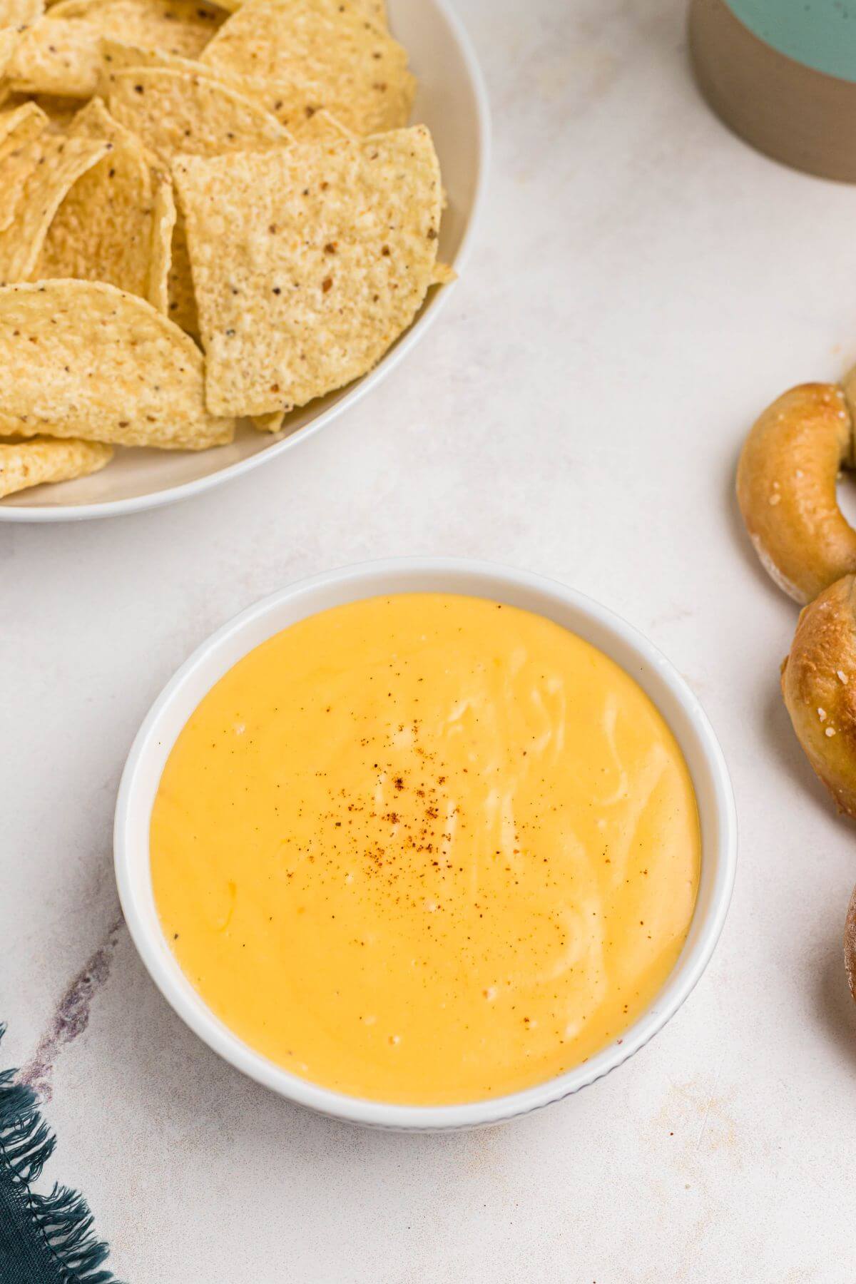 Creamy cheese sauce in a white bowl with chips and soft pretzels on the table. 