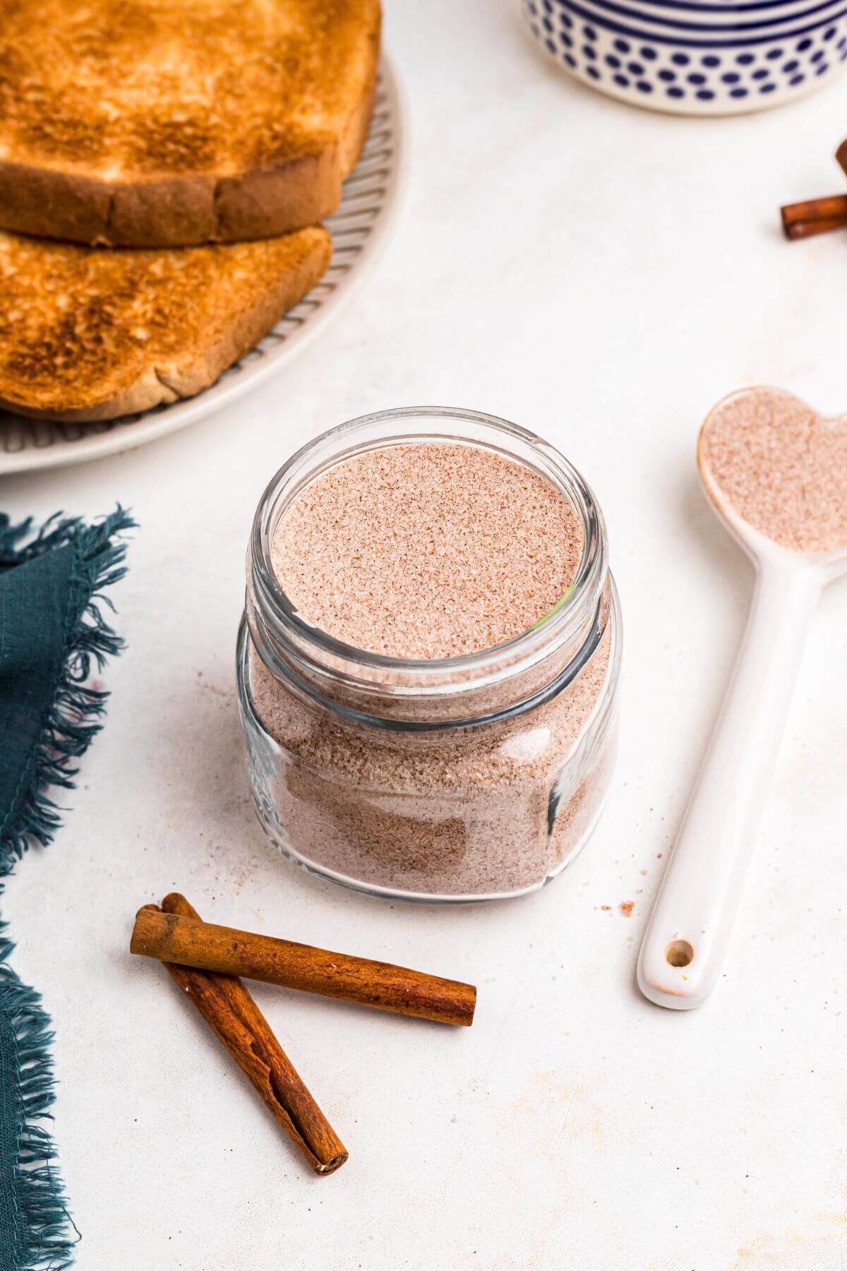 Cinnamon sugar mixture being stored in a clear glass jar in front of toast on a marble table. 