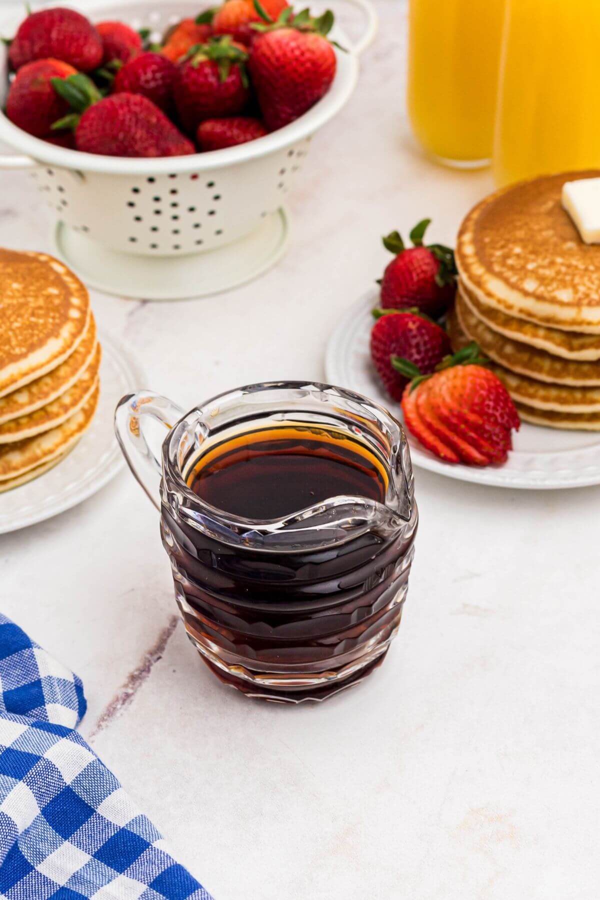 Small glass serving pitcher filled with homemade syrup in front of plates with stacks of pancakes and strawberries. 